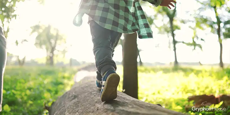 Young child shown from the waist down walking along the top of a log in the woods