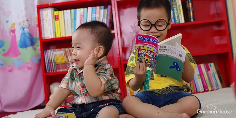 Two toddler children looking at a book while sitting on the floor, with a bookcase behind them